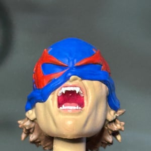 May include: A miniature sculpted head featuring a blue mask with red accents covering the eyes. The mouth is wide open in a scream, revealing white fangs and a red interior. The figure has brown hair visible at the base of the neck and a tan skin tone.