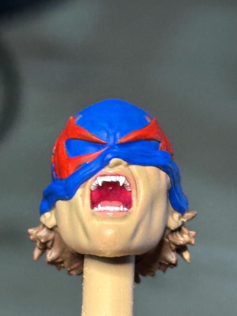 May include: A miniature sculpted head featuring a blue mask with red accents covering the eyes. The mouth is wide open in a scream, revealing white fangs and a red interior. The figure has brown hair visible at the base of the neck and a tan skin tone.