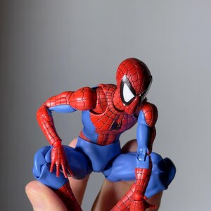 Puede incluir: Una figura de acci&oacute;n de Spider-Man en posici&oacute;n agachada. La figura es predominantemente roja y azul, con un patr&oacute;n de telara&ntilde;a detallado en las secciones rojas. Los ojos de la figura son blancos y se sostiene en una mano.
