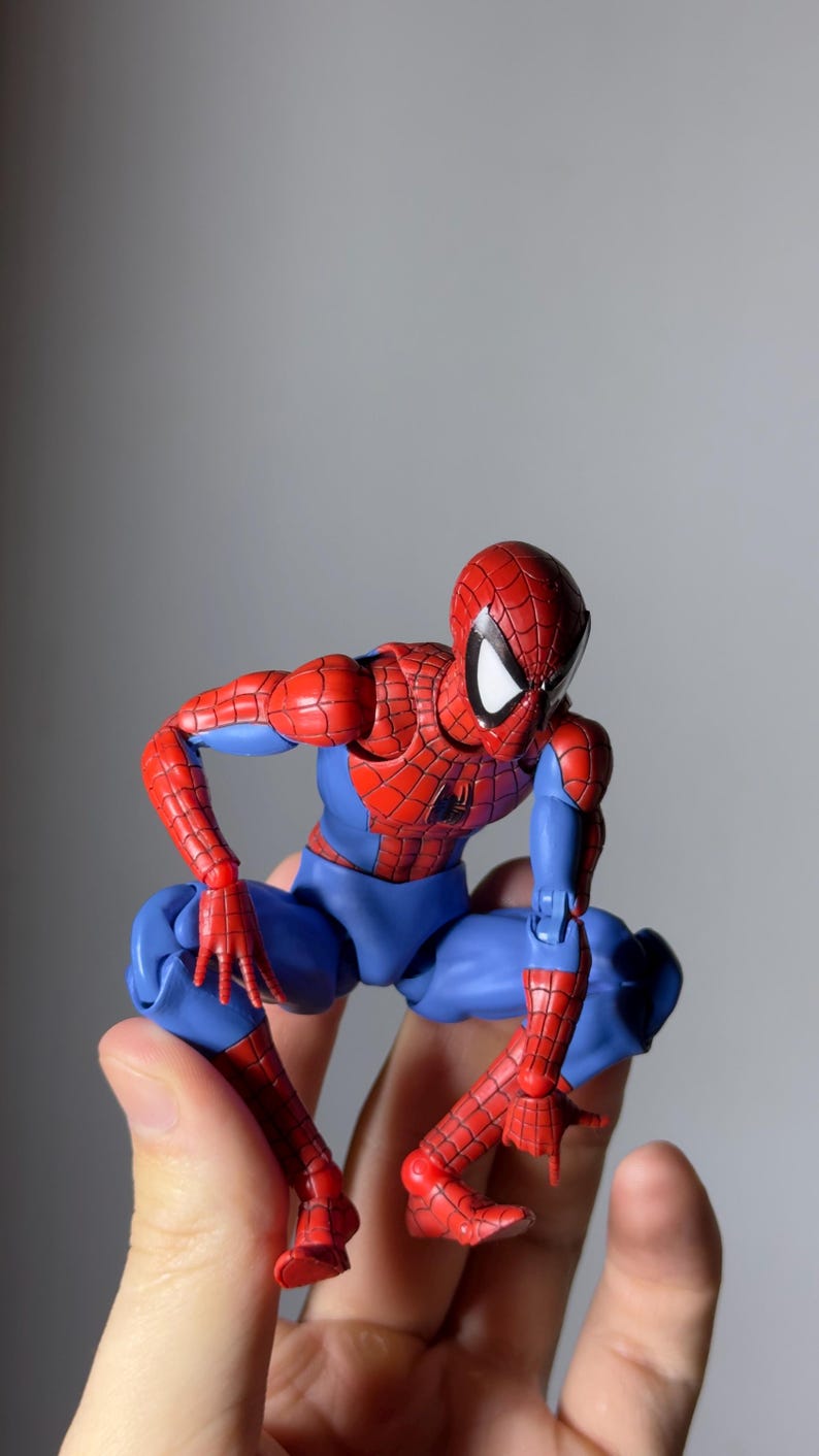 Puede incluir: Una figura de acci&oacute;n de Spider-Man en posici&oacute;n agachada. La figura es predominantemente roja y azul, con un patr&oacute;n de telara&ntilde;a detallado en las secciones rojas. Los ojos de la figura son blancos y se sostiene en una mano.