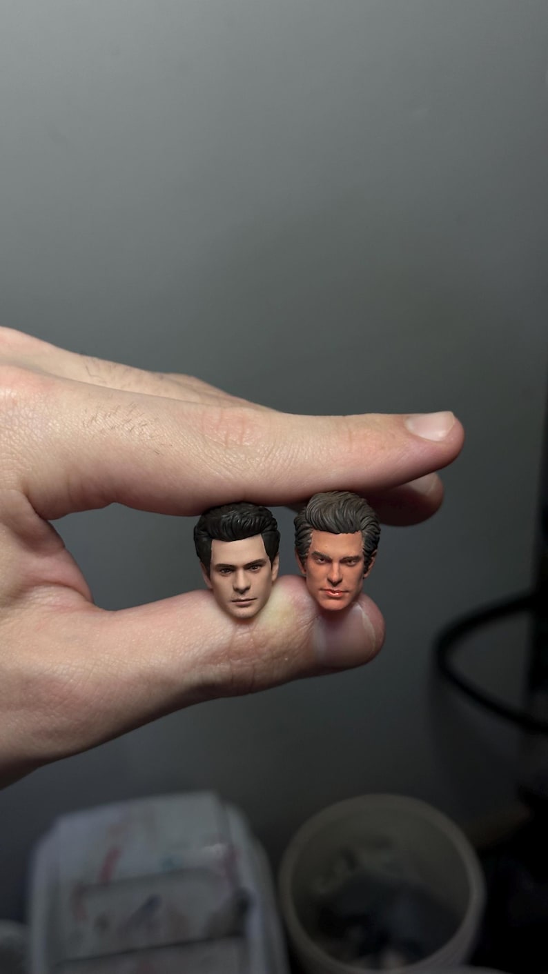 Może przedstawiać: Dwie szczeg&oacute;łowe miniaturowe rzeźby głowy, jedna z ciemnymi włosami, a druga z jasnobrązowymi włosami. Głowy trzymane są między palcami na szarym tle. Rzeźby wydają się być realistycznymi przedstawieniami osoby.