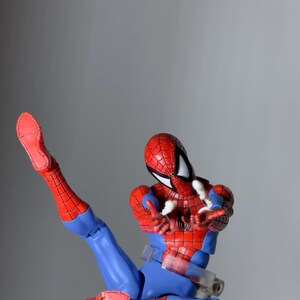 Puede incluir: Figura de acci&oacute;n de Spider-Man en una pose din&aacute;mica, con un traje rojo y azul con detalles de telara&ntilde;a. La figura est&aacute; en plena acci&oacute;n, con una pierna extendida y los brazos extendidos, sobre un fondo gris neutro.