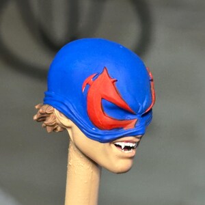May include: A sculpted figurine head featuring a bright blue mask with prominent red eye-shaped markings. The face has a light skin tone, visible brown hair at the back, and an open mouth revealing white teeth with sharp, pointed fangs.