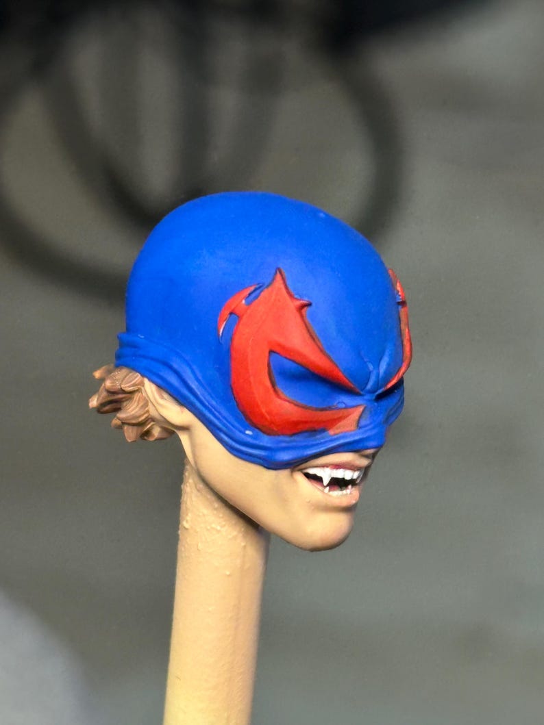May include: A sculpted figurine head featuring a bright blue mask with prominent red eye-shaped markings. The face has a light skin tone, visible brown hair at the back, and an open mouth revealing white teeth with sharp, pointed fangs.