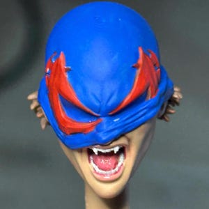 May include: A sculpted head figure featuring a bright blue mask with jagged red eye markings covering the upper face. The mouth is open in a snarl, revealing white fangs and a pink tongue. Brown hair is visible on the sides of the head.