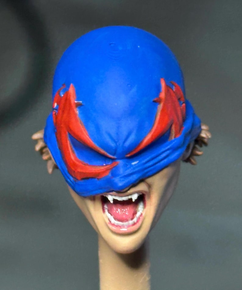 May include: A sculpted head figure featuring a bright blue mask with jagged red eye markings covering the upper face. The mouth is open in a snarl, revealing white fangs and a pink tongue. Brown hair is visible on the sides of the head.