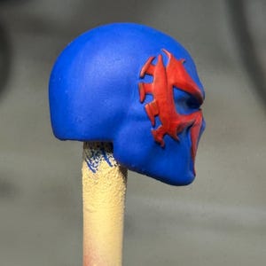 May include: A blue helmet or mask accessory featuring a prominent, raised red spider-like emblem on the side. The object is mounted on a cylindrical, textured beige stick, showcasing a smooth, matte finish and a distinct, stylized facial design.