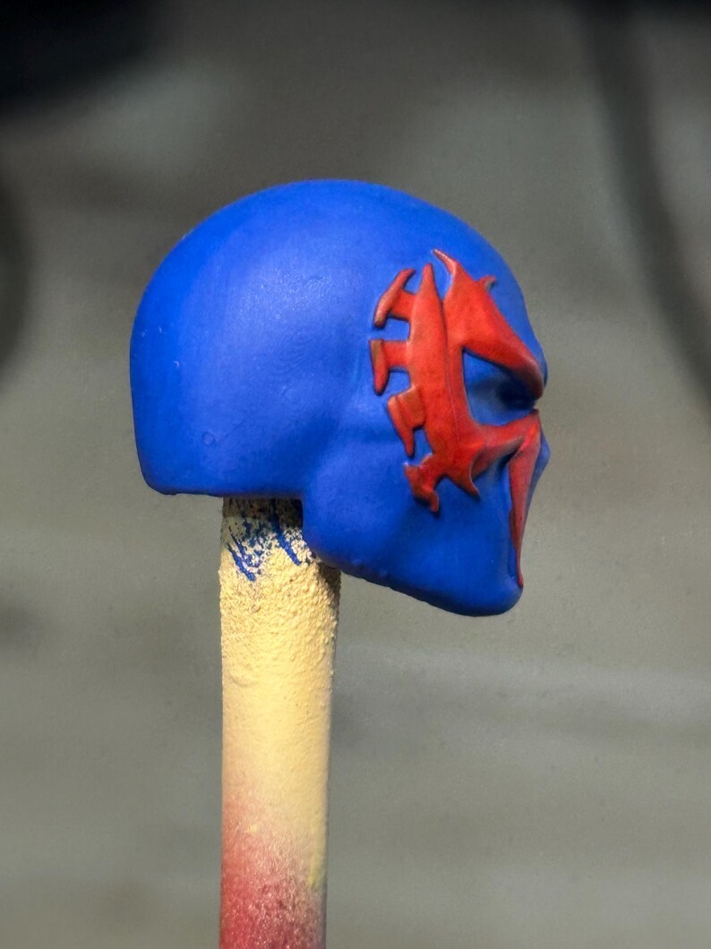 May include: A blue helmet or mask accessory featuring a prominent, raised red spider-like emblem on the side. The object is mounted on a cylindrical, textured beige stick, showcasing a smooth, matte finish and a distinct, stylized facial design.