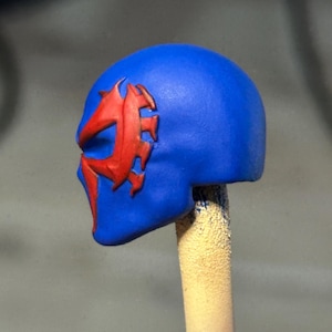 May include: A blue sculpted head piece featuring a raised, jagged red emblem on the side. The object is mounted on a cylindrical stick, displaying a smooth matte finish with distinct contours around the eye and cheek area of the mask design.