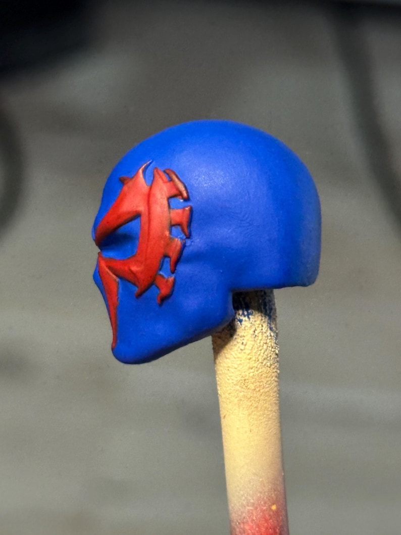 May include: A blue sculpted head piece featuring a raised, jagged red emblem on the side. The object is mounted on a cylindrical stick, displaying a smooth matte finish with distinct contours around the eye and cheek area of the mask design.