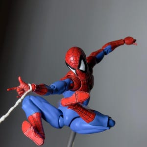 Puede incluir: Figura de acci&oacute;n de Spider-Man en pose din&aacute;mica, con un traje rojo y azul con detalles de telara&ntilde;a. La figura est&aacute; en el aire, con una mano extendida y un hilo blanco. La figura est&aacute; sostenida por un soporte transparente.