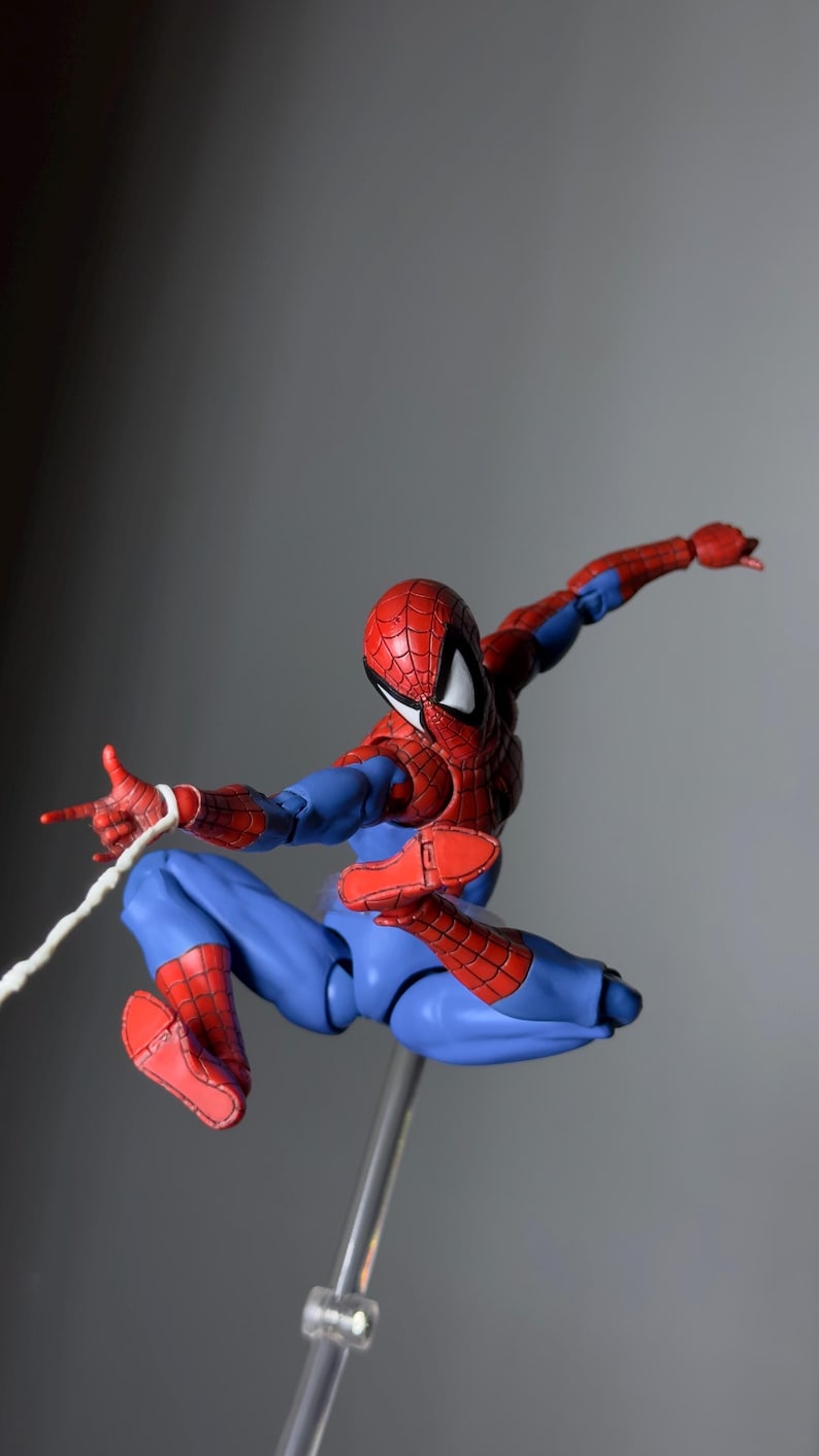 Puede incluir: Figura de acci&oacute;n de Spider-Man en pose din&aacute;mica, con un traje rojo y azul con detalles de telara&ntilde;a. La figura est&aacute; en el aire, con una mano extendida y un hilo blanco. La figura est&aacute; sostenida por un soporte transparente.