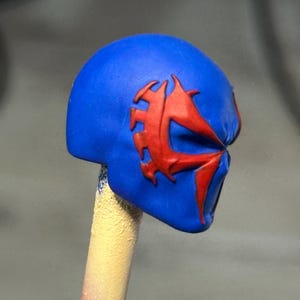 May include: A blue miniature helmet head sculpt featuring a prominent red spider-like emblem embossed on the side. The head is mounted on a thin, cylindrical stick that transitions from a yellow to a red color at the base, providing support.