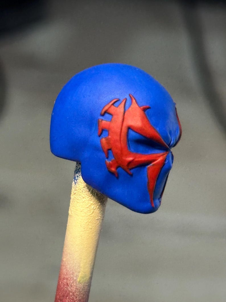 May include: A blue miniature helmet head sculpt featuring a prominent red spider-like emblem embossed on the side. The head is mounted on a thin, cylindrical stick that transitions from a yellow to a red color at the base, providing support.