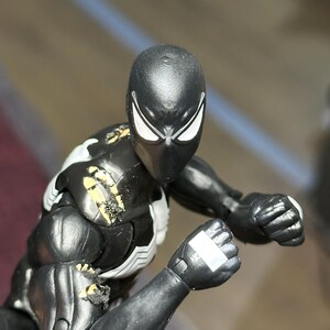 May include: A black action figure with white eye lenses and a white chest emblem. The figure features visible paint wear on the right shoulder and upper arm, revealing yellow plastic underneath the black surface. The figure is posed with clenched fists.