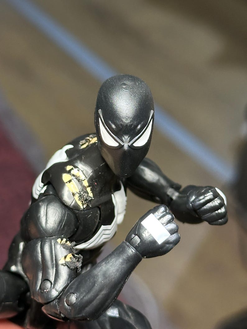May include: A black action figure with white eye lenses and a white chest emblem. The figure features visible paint wear on the right shoulder and upper arm, revealing yellow plastic underneath the black surface. The figure is posed with clenched fists.