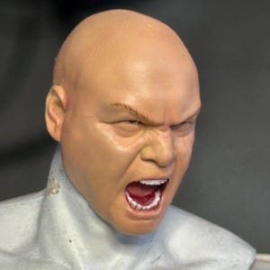 Könnte beinhalten: Ein skulptierter Figuren-Kopf eines kahlen, hellhäutigen Charakters mit einem grimmigen, schreienden Gesichtsausdruck, sichtbaren Zähnen und gerunzelter Stirn. Die Figur hat einen glatten, hautfarbenen Teint und ist an einem hellgrauen Torso befestigt.