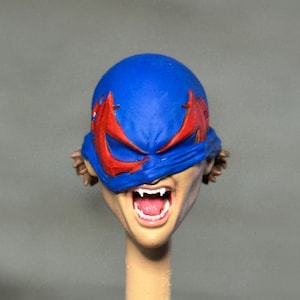 May include: A painted miniature head featuring a blue mask with red eye accents covering the upper face. The mouth is open in a wide expression, revealing white fangs and a pink tongue. Brown hair is visible at the sides of the head.
