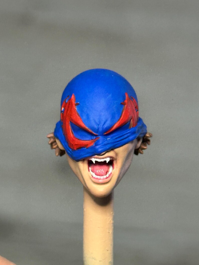 May include: A painted miniature head featuring a blue mask with red eye accents covering the upper face. The mouth is open in a wide expression, revealing white fangs and a pink tongue. Brown hair is visible at the sides of the head.