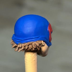 May include: A miniature figurine head featuring a bright blue helmet with a red emblem on the side. The head has sculpted brown hair visible beneath the helmet edge and is mounted on a light-colored cylindrical stick for painting and display purposes.