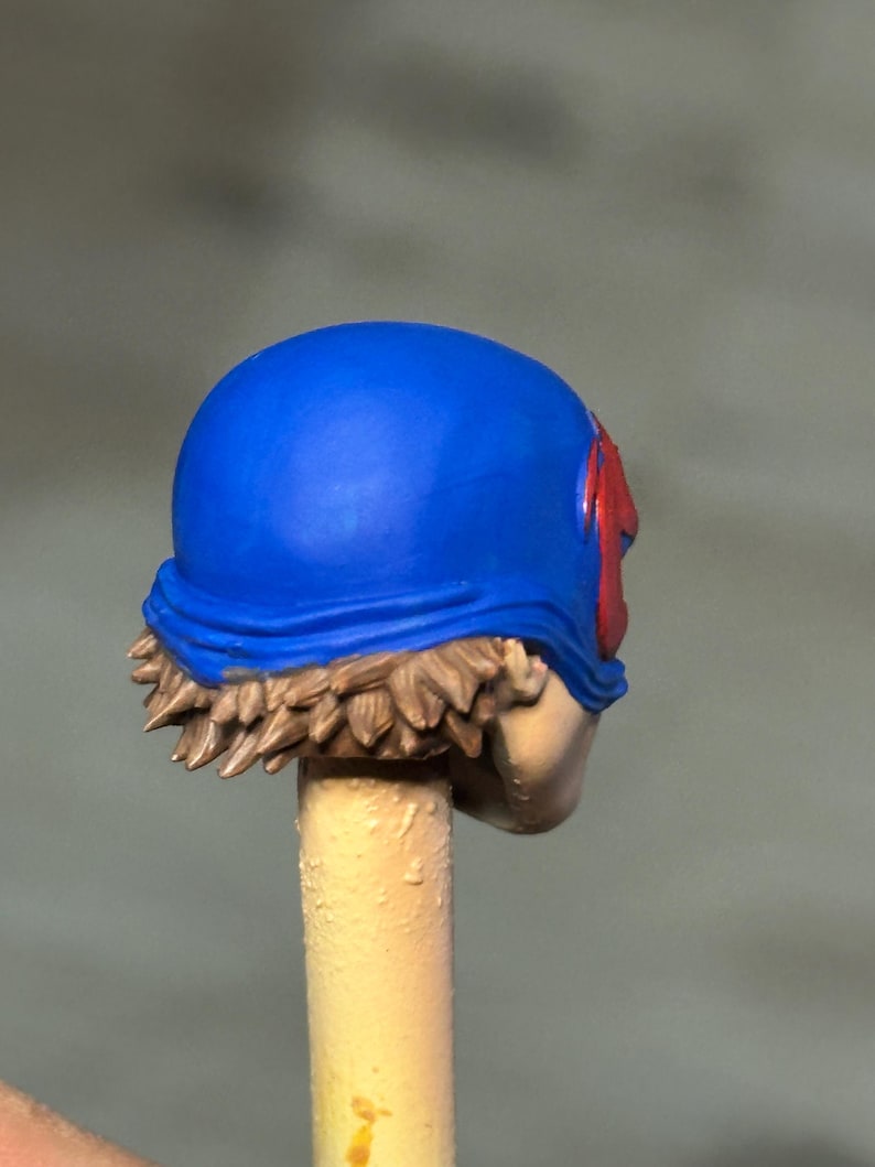 May include: A miniature figurine head featuring a bright blue helmet with a red emblem on the side. The head has sculpted brown hair visible beneath the helmet edge and is mounted on a light-colored cylindrical stick for painting and display purposes.