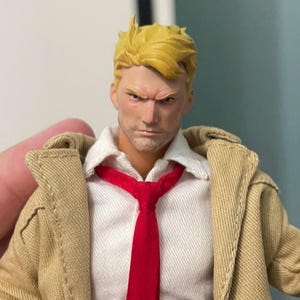 May include: A collectible action figure featuring a character with short blonde hair, a stern facial expression, a white collared shirt, a bright red necktie, and a textured tan trench coat. The figure is rendered with realistic facial features.