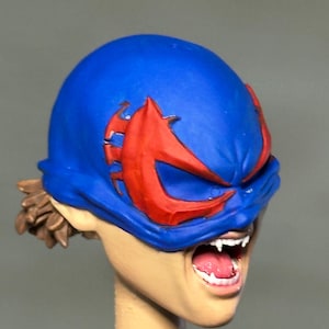 May include: A miniature sculpted head featuring a bright blue mask with large red eye cutouts. The figure has an open mouth revealing white teeth and fangs, with brown hair visible at the back. The skin tone is light beige, and the expression is intense.