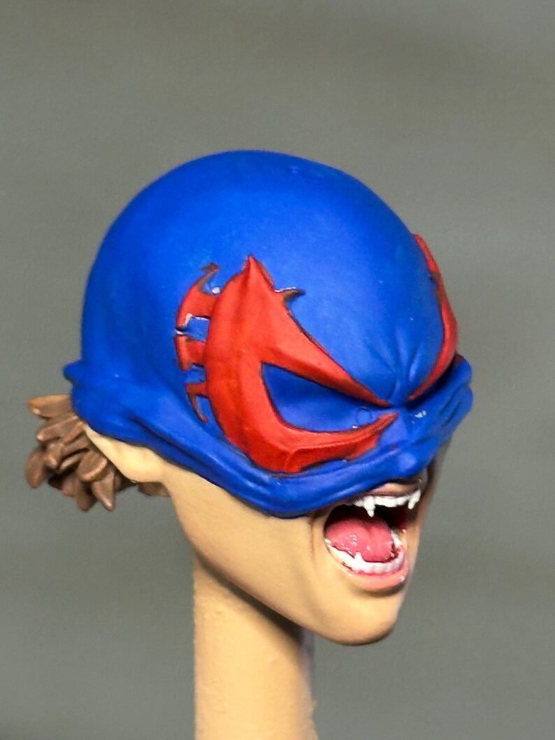May include: A miniature sculpted head featuring a bright blue mask with large red eye cutouts. The figure has an open mouth revealing white teeth and fangs, with brown hair visible at the back. The skin tone is light beige, and the expression is intense.
