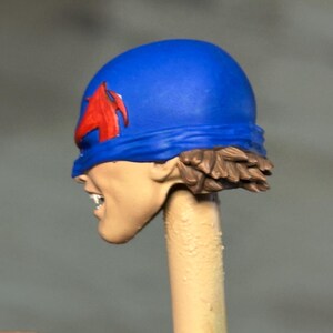 May include: A painted miniature head featuring a bright blue cowl with a red emblem on the side. The figure has light skin, visible white teeth, and short brown hair at the base of the cowl. The head is mounted on a thin, light-colored cylindrical post.