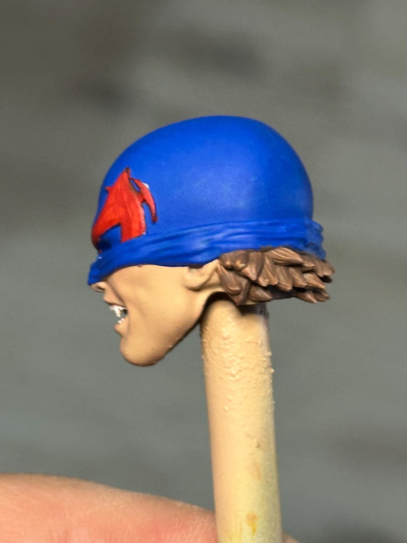 May include: A painted miniature head featuring a bright blue cowl with a red emblem on the side. The figure has light skin, visible white teeth, and short brown hair at the base of the cowl. The head is mounted on a thin, light-colored cylindrical post.