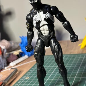 May include: A black action figure with a white spider emblem on the chest and white eye patches. The figure has articulated joints, a muscular build, and visible wear on the right shoulder and thigh. It stands on a green cutting mat with grid lines.