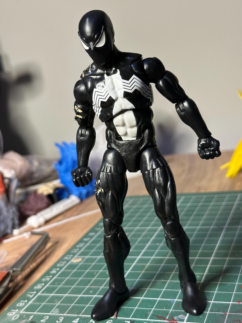 May include: A black action figure with a white spider emblem on the chest and white eye patches. The figure has articulated joints, a muscular build, and visible wear on the right shoulder and thigh. It stands on a green cutting mat with grid lines.
