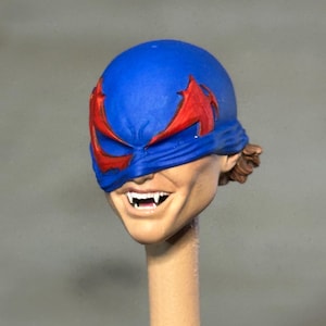 May include: A sculpted action figure head featuring a blue mask with red eye accents. The face has a wide, toothy grin revealing sharp, pointed fangs. The skin tone is light, and a small amount of brown hair is visible at the back of the head.