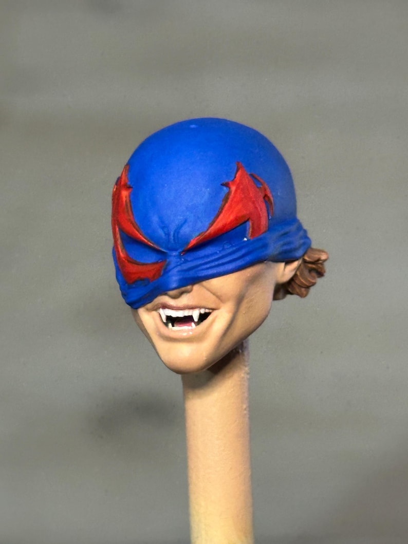 May include: A sculpted action figure head featuring a blue mask with red eye accents. The face has a wide, toothy grin revealing sharp, pointed fangs. The skin tone is light, and a small amount of brown hair is visible at the back of the head.