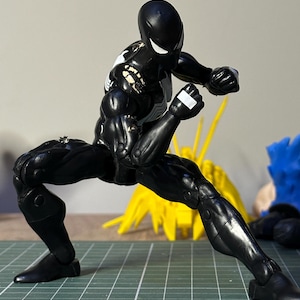 May include: A black action figure in a dynamic fighting pose, featuring a white spider emblem on the chest and back. The figure has white eyes and visible wear on the shoulders. It is positioned on a green cutting mat with inch markings.
