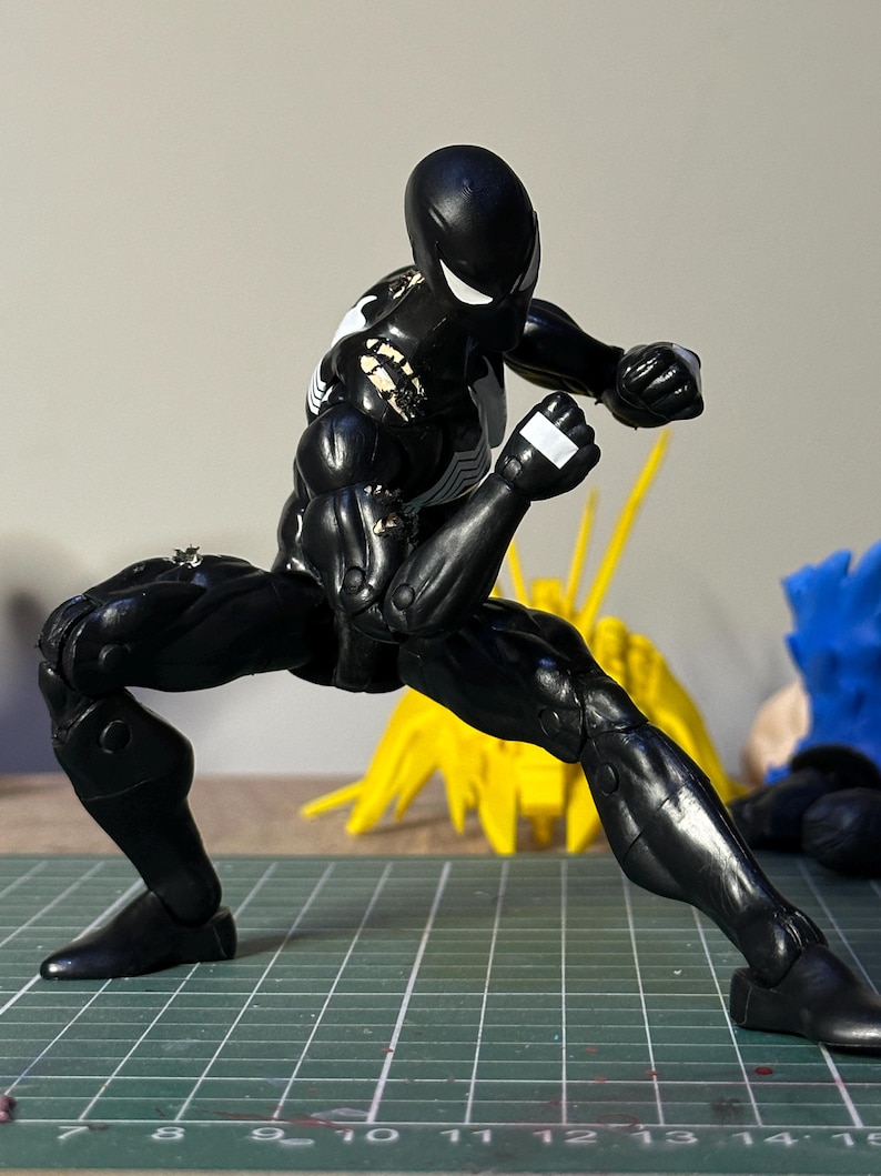 May include: A black action figure in a dynamic fighting pose, featuring a white spider emblem on the chest and back. The figure has white eyes and visible wear on the shoulders. It is positioned on a green cutting mat with inch markings.