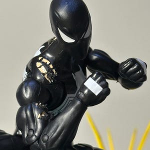 May include: A black action figure with white eye lenses and a white spider emblem on the chest. The figure is in a dynamic fighting pose with clenched fists, featuring visible wear and paint chips on the right shoulder and arm area.
