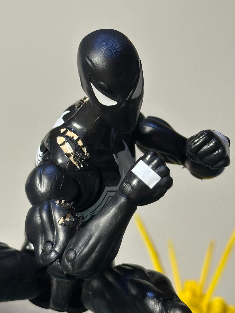 May include: A black action figure with white eye lenses and a white spider emblem on the chest. The figure is in a dynamic fighting pose with clenched fists, featuring visible wear and paint chips on the right shoulder and arm area.