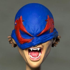 May include: A sculpted action figure head featuring a blue mask with red eye accents covering the upper face. The mouth is open in a wide expression, revealing white fangs and a pink tongue. Brown hair is visible on the sides of the head.