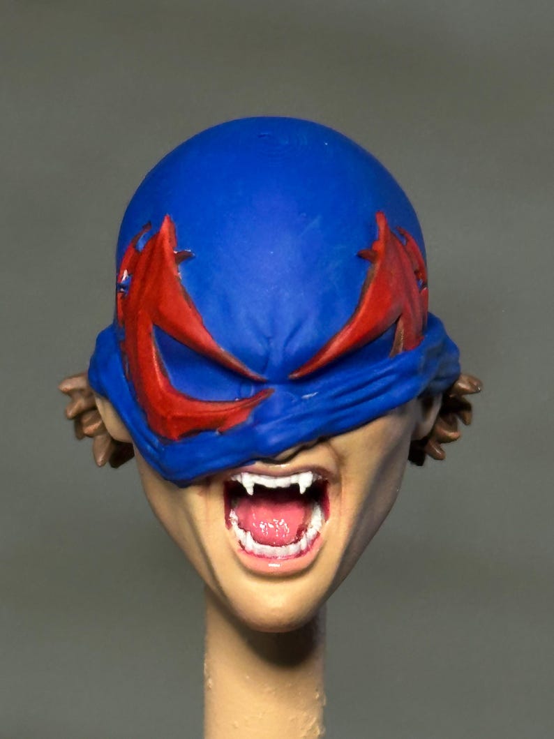 May include: A sculpted action figure head featuring a blue mask with red eye accents covering the upper face. The mouth is open in a wide expression, revealing white fangs and a pink tongue. Brown hair is visible on the sides of the head.
