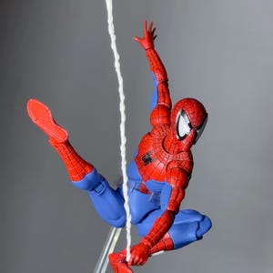 Puede incluir: Figura de acci&oacute;n de Spider-Man, rojo y azul, en pose din&aacute;mica, suspendida en el aire por una telara&ntilde;a blanca. La figura tiene una m&aacute;scara, traje y botas rojas, contrastadas con guantes y pantalones azules. Soporte transparente.