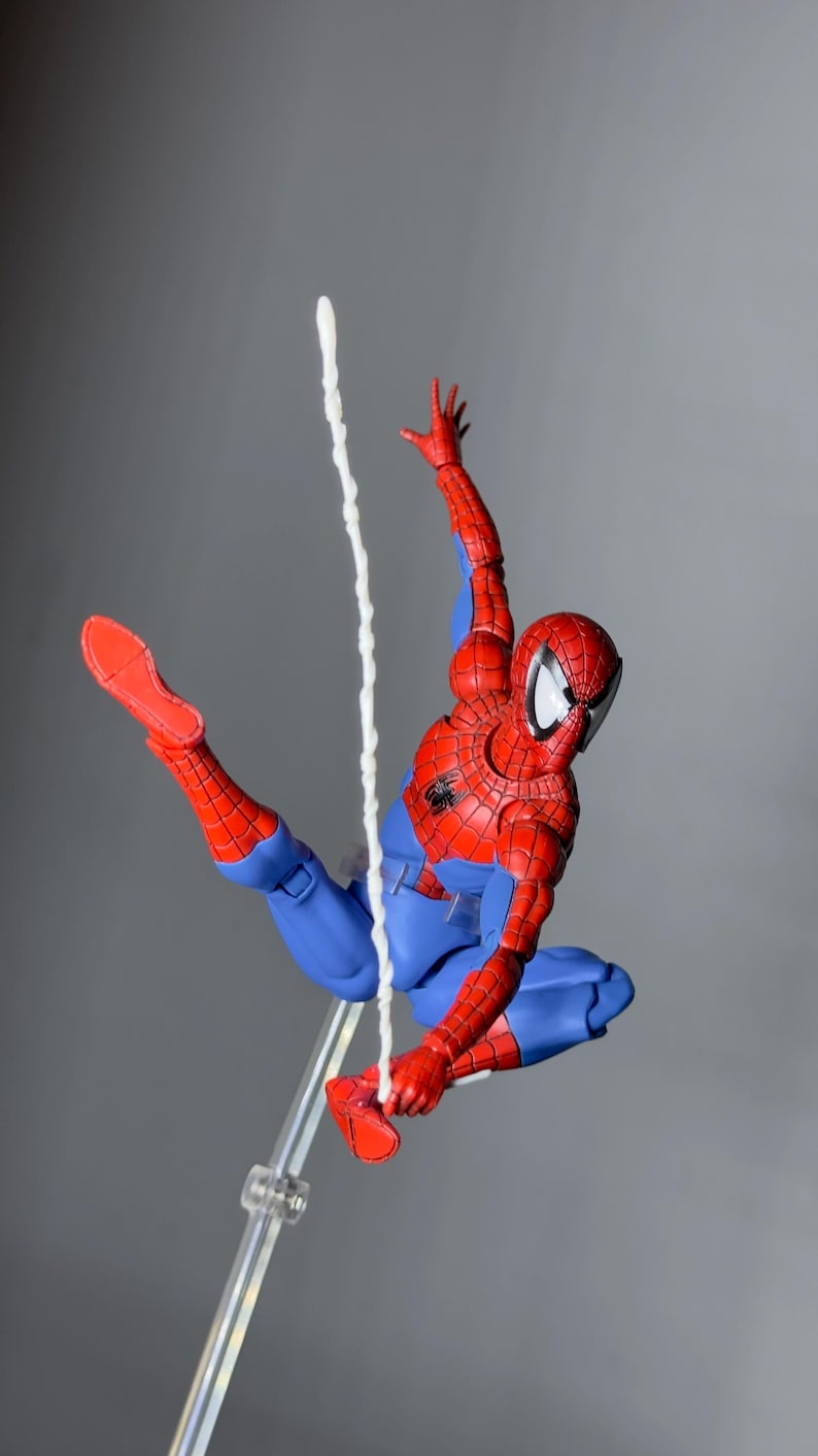 Puede incluir: Figura de acci&oacute;n de Spider-Man, rojo y azul, en pose din&aacute;mica, suspendida en el aire por una telara&ntilde;a blanca. La figura tiene una m&aacute;scara, traje y botas rojas, contrastadas con guantes y pantalones azules. Soporte transparente.