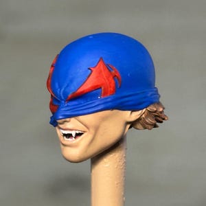 May include: A sculpted action figure head featuring a blue mask with red wing-like accents covering the eyes. The face has a light skin tone, a wide grin revealing white fangs, and short brown hair visible at the back of the neck.