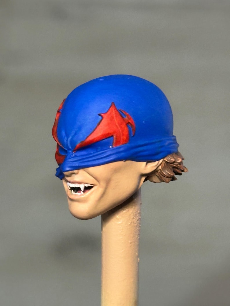 May include: A sculpted action figure head featuring a blue mask with red wing-like accents covering the eyes. The face has a light skin tone, a wide grin revealing white fangs, and short brown hair visible at the back of the neck.