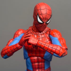 May include: A red and blue Spider-Man action figure in a dynamic pose. The figure features a detailed web pattern on the red sections of the suit, with blue accents on the arms and torso. The figure's hands are clasped together.