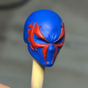 May include: A blue sculpted head piece featuring a prominent red spider-like emblem across the eye area. The mask has a smooth, matte finish with sharp, angular lines defining the facial features and the stylized red markings extending to the temples.