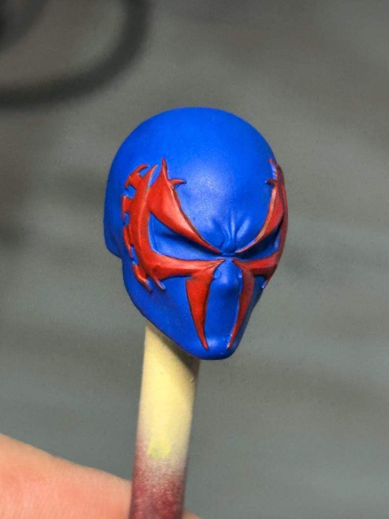 May include: A blue sculpted head piece featuring a prominent red spider-like emblem across the eye area. The mask has a smooth, matte finish with sharp, angular lines defining the facial features and the stylized red markings extending to the temples.