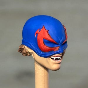 May include: A sculpted action figure head featuring a bright blue mask with prominent red eye-shaped accents. The face has a light skin tone, visible brown hair at the back, and a wide grin revealing sharp white fangs, mounted on a beige post.