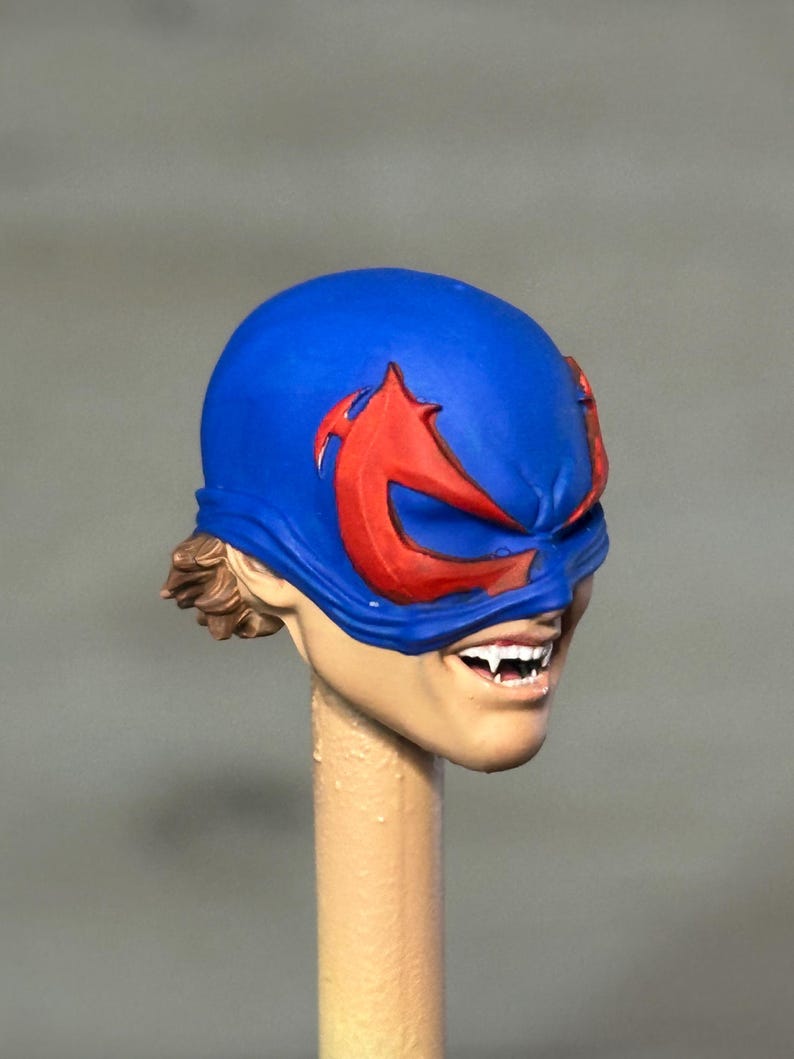 May include: A sculpted action figure head featuring a bright blue mask with prominent red eye-shaped accents. The face has a light skin tone, visible brown hair at the back, and a wide grin revealing sharp white fangs, mounted on a beige post.