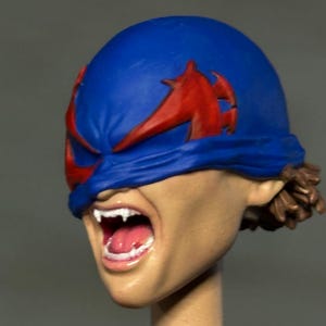 May include: A sculpted head model featuring a blue mask with red wing-like accents covering the eyes. The mouth is open in a wide expression, revealing white fangs and a pink tongue. The skin tone is light tan, and brown hair is visible at the back.